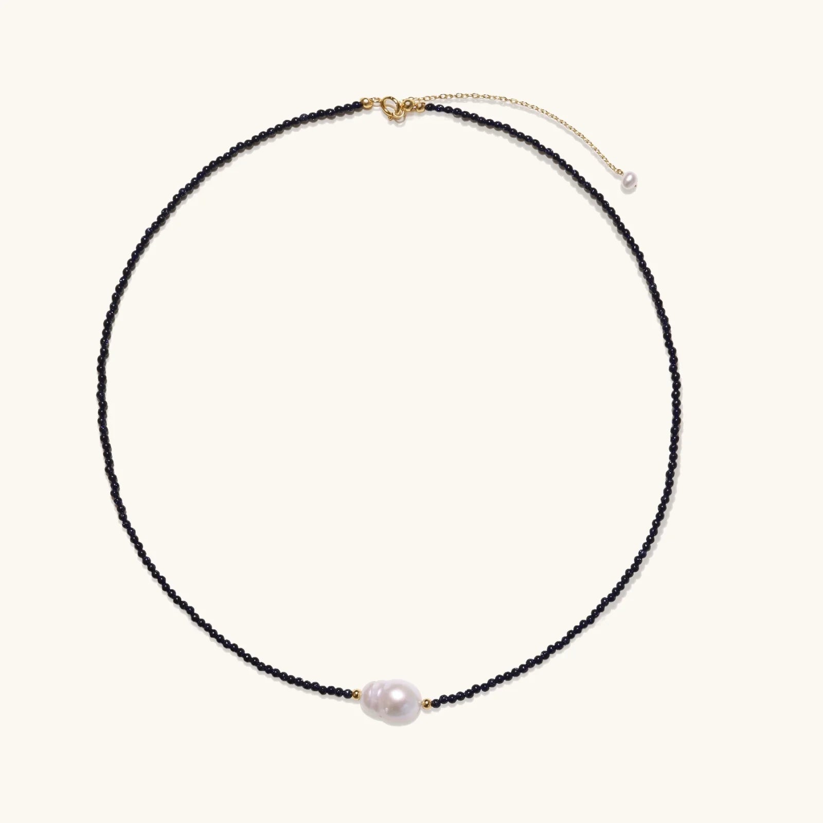 Delicate necklace with black blue sandstone beads and baroque pearl center on white background with gold accents.