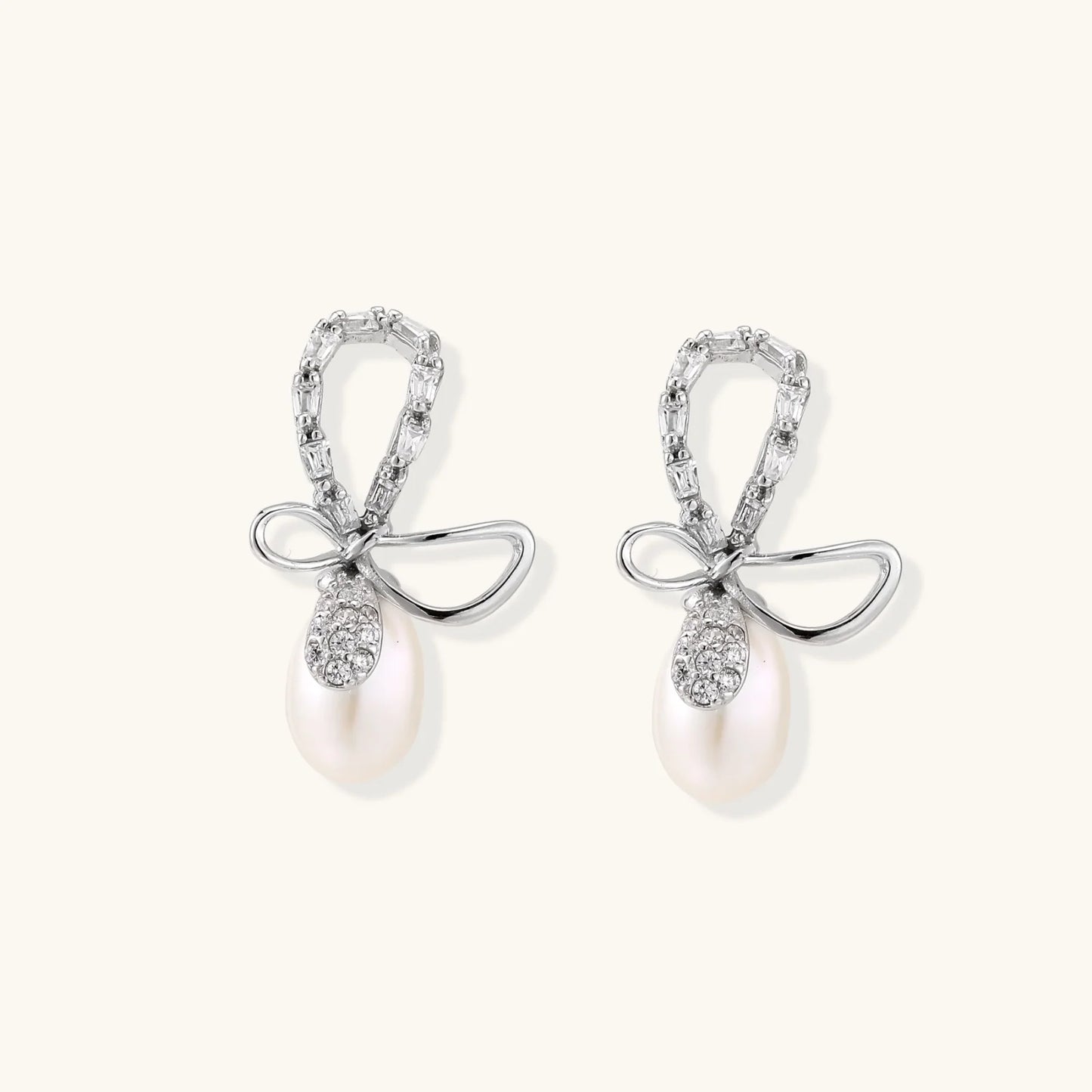 Silver bow-shaped stud earrings with baguette crystals and dangling white freshwater pearls on cream background