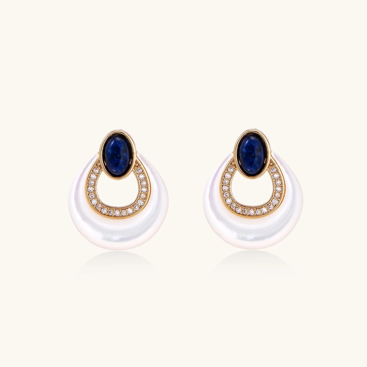 Gold drop earrings with oval lapis lazuli, crystal-paved frame, and iridescent mother-of-pearl teardrop on cream background