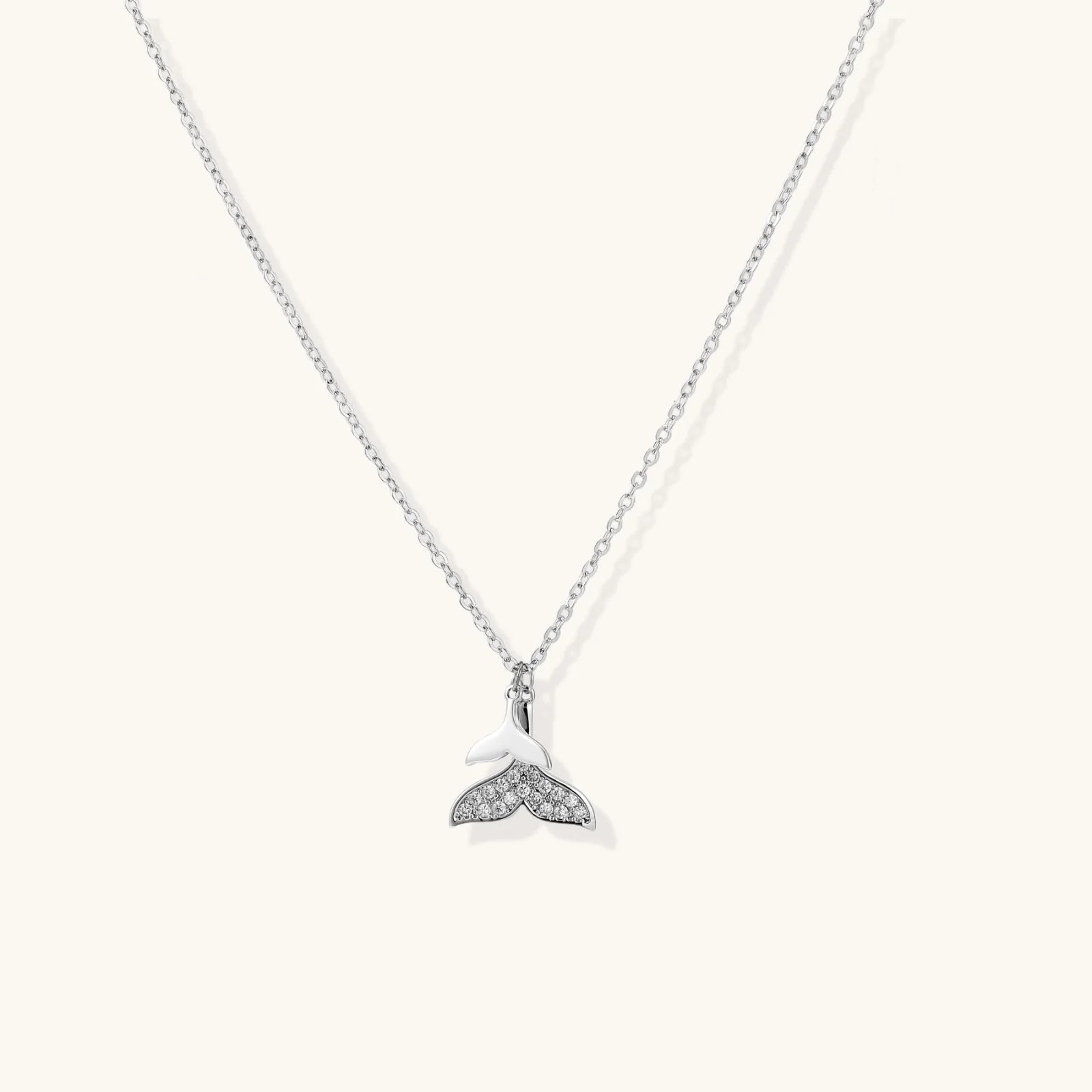 Delicate white gold plated whale tail pendant with pavé crystals on silver chain against cream background.