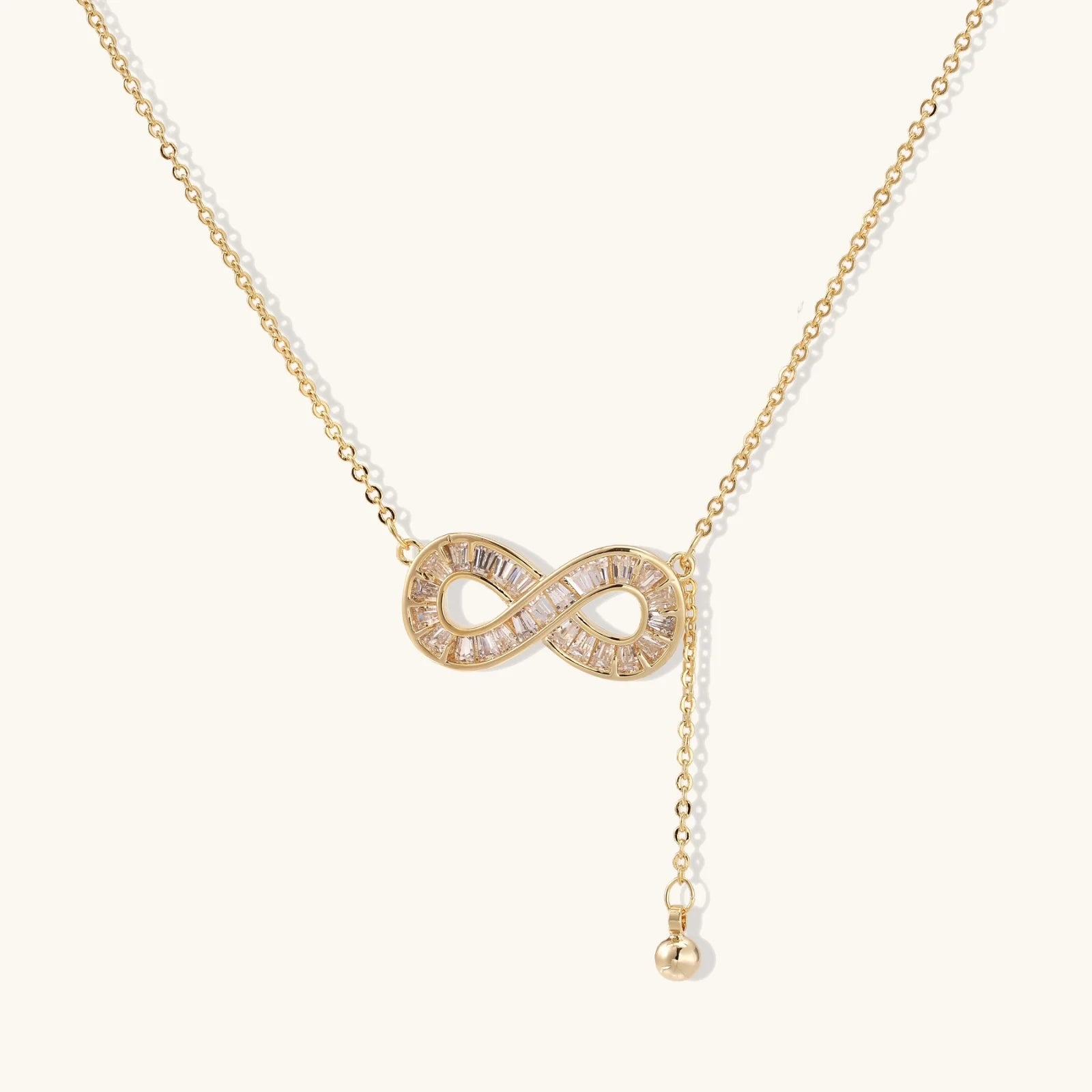 Gold infinity pendant with baguette crystals on adjustable chain against cream background.